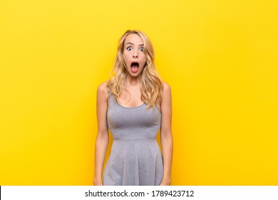 young blonde woman looking very shocked surprised  staring and open mouth saying wow against orange wall