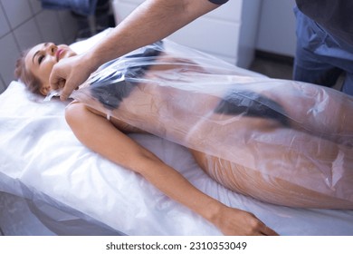Young blonde woman makes cosmetology procedure in spa salon. Full anti cellulite body wrap. Male beautician covers his female client with plastic wrap. Modern beauty salon interior. Copy space. - Shutterstock ID 2310353049