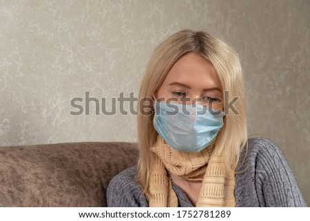 young blonde woman in a medical mask with a scarf around her neck sits sad on the sofa with downcast eyes, copy space