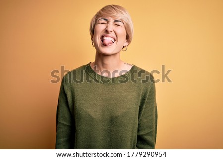Young blonde woman with modern short hair wearing casual sweater over yellow background sticking tongue out happy with funny expression. Emotion concept.