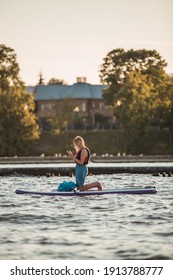 A Young Blonde Woman On The Phone On A SUP Stand Up Paddle Board In The River In Summer, Golden Hour