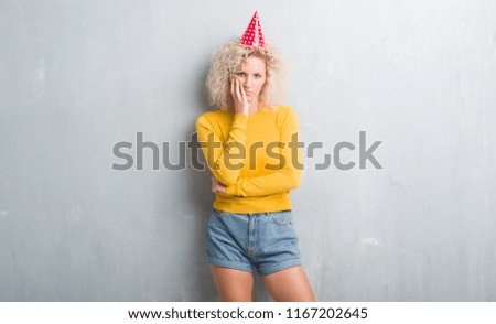 Young blonde woman over grunge grey wall wearing birthday hat thinking looking tired and bored with depression problems with crossed arms.