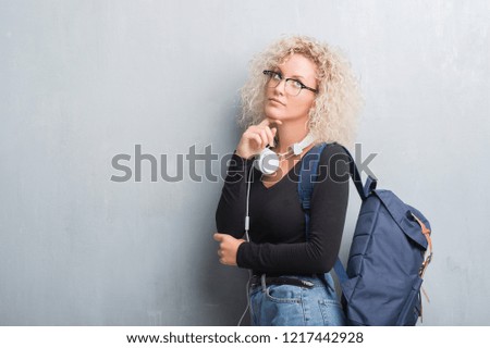 Young blonde woman over grunge grey wall wearing backpack and headphones with hand on chin thinking about question, pensive expression. Smiling with thoughtful face. Doubt concept.