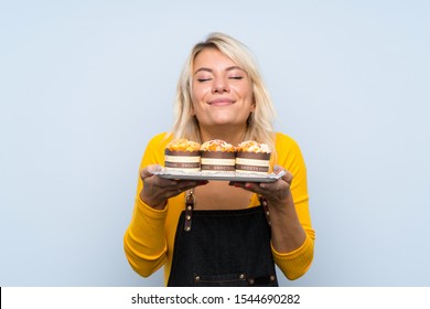 Young blonde woman over isolated background holding mini cakes enjoying the smell of them