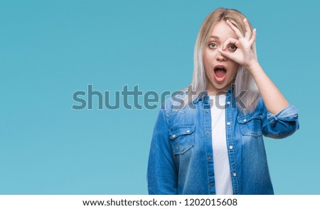 Young blonde woman over isolated background doing ok gesture shocked with surprised face, eye looking through fingers. Unbelieving expression.
