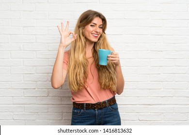 Young blonde woman over white brick wall holding hot cup of coffee