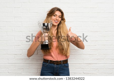 Young blonde woman over white brick wall holding a trophy