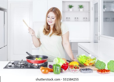 Young Blonde Woman With Overweight Body, Cooking Vegetables In The Kitchen At Home