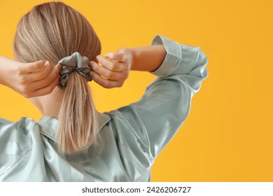 Young blonde woman in pajamas doing ponytail with scrunchy on yellow background, back view - Powered by Shutterstock