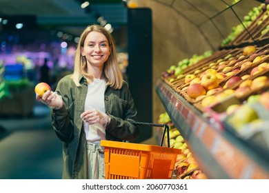 young blonde woman picks chooses fruits vegetables on the counter in supermarket. Female housewife shopping in market standing near vegetable department store with a basket in hands. Examines apple - Powered by Shutterstock