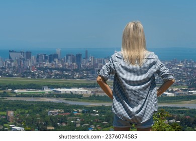 Young blonde woman in a plaid shirt looks at the city from the height of the mountain from the back. Girl with blonde hair looks down at the city, the sea. Concept of travel and wanderlust. - Powered by Shutterstock