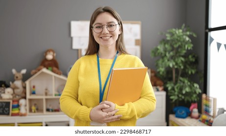 Young blonde woman preschool teacher smiling confident holding books at kindergarten - Powered by Shutterstock