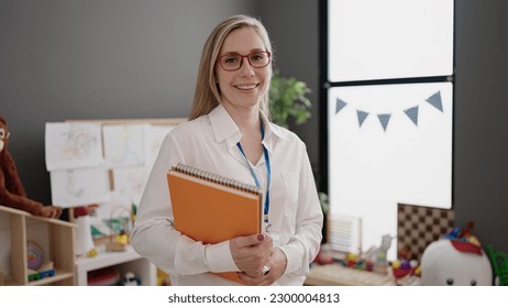 Young blonde woman preschool teacher smiling confident holding books at kindergarten - Powered by Shutterstock