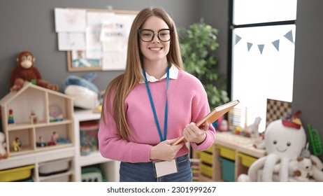 Young blonde woman preschool teacher smiling confident holding book at kindergarten - Powered by Shutterstock