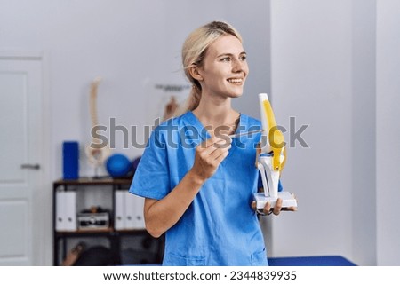 Young blonde woman pysiotherapist smiling confident pointing to anatomical model of knee at rehab clinic