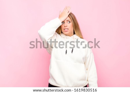 young blonde woman raising palm to forehead thinking oops, after making a stupid mistake or remembering, feeling dumb against flat wall