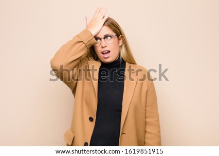 young blonde woman raising palm to forehead thinking oops, after making a stupid mistake or remembering, feeling dumb against flat wall