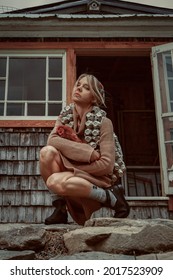 Young Blonde Woman With A Scarf On Her Head, Wearing A Knitted Dress And A Down Vest Made Of Egg Cartons, Holding A Brown Hen In Her Arms, Near A Chicken Coop