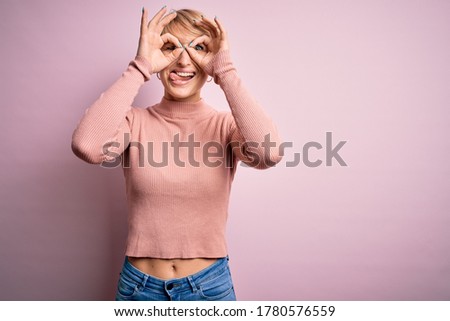 Young blonde woman with short hair wearing casual turtleneck sweater over pink background doing ok gesture like binoculars sticking tongue out, eyes looking through fingers. Crazy expression.