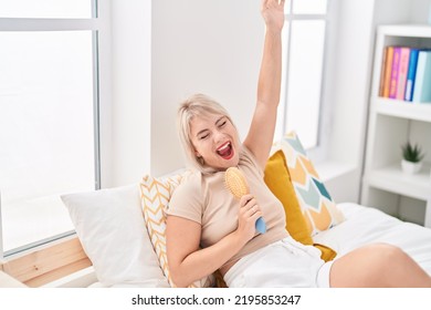 Young Blonde Woman Singing Song Using Brush As A Microphone At Bedroom