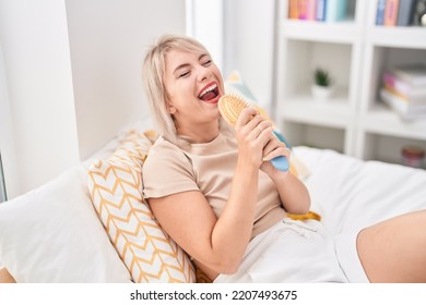 Young Blonde Woman Singing Song Using Brush As A Microphone At Bedroom