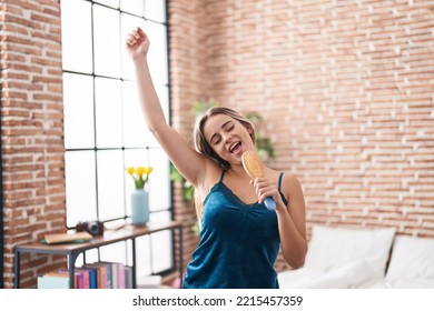 Young Blonde Woman Singing Song Using Brush As A Microphone At Bedroom