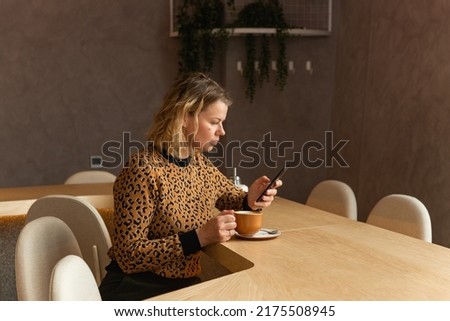 a young blonde woman is sitting in a cafe in beige tones, drinking coffee, scrolling on the phone, and smiling