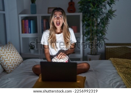 Young blonde woman sitting on the bed at home watching a movie from laptop angry and mad screaming frustrated and furious, shouting with anger looking up. 