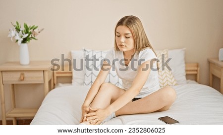Young blonde woman sitting on bed massaging leg at bedroom