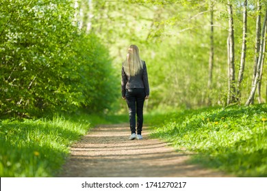 Young Blonde Woman Slowly Walking On Natural Trail In Green Forest. Warm Sunny Spring Day. Spending Time Alone In Nature. Peaceful Atmosphere. Back View.  