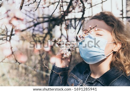 Young blonde woman smells cherry tree blossom through medical mask. Conceptual image of corona virus quarantine and allergy.