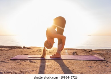 Young blonde woman in sportswear doing yoga asanas Uttanasana pose on the seashore at sunrise on purple yoga mat. Sports, meditation by the ocean on the yellow sand. Positive effects on brain function