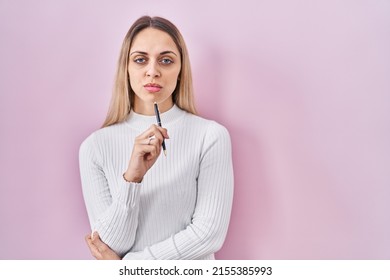 Young Blonde Woman Standing Holding Pen Over Pink Background Skeptic And Nervous, Frowning Upset Because Of Problem. Negative Person. 