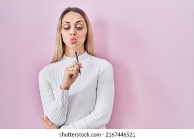 Young Blonde Woman Standing Holding Pen Over Pink Background Making Fish Face With Mouth And Squinting Eyes, Crazy And Comical. 