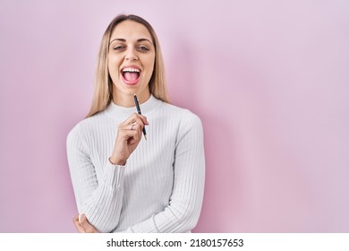 Young Blonde Woman Standing Holding Pen Over Pink Background Smiling And Laughing Hard Out Loud Because Funny Crazy Joke. 