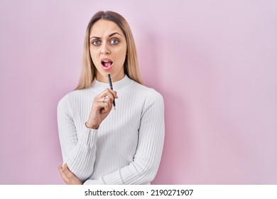 Young Blonde Woman Standing Holding Pen Over Pink Background Clueless And Confused Expression. Doubt Concept. 