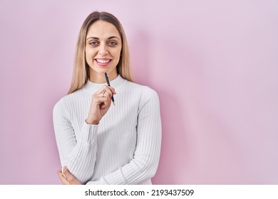 Young Blonde Woman Standing Holding Pen Over Pink Background Smiling With A Happy And Cool Smile On Face. Showing Teeth. 