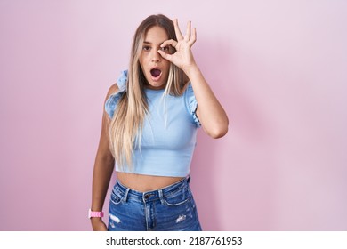 Young blonde woman standing over pink background doing ok gesture shocked with surprised face, eye looking through fingers. unbelieving expression. 
