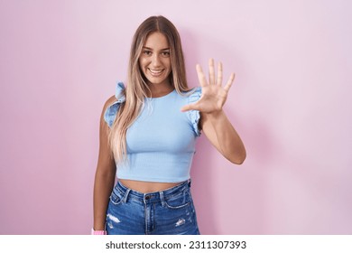 Young blonde woman standing over pink background showing and pointing up with fingers number five while smiling confident and happy.  - Shutterstock ID 2311307393