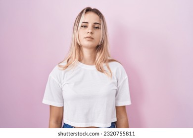 Young blonde woman standing over pink background relaxed with serious expression on face. simple and natural looking at the camera. 