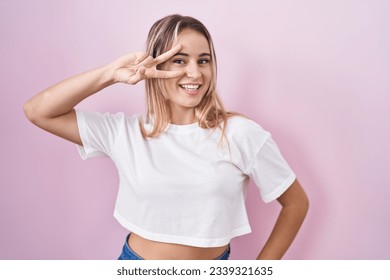 Young blonde woman standing over pink background doing peace symbol with fingers over face, smiling cheerful showing victory 