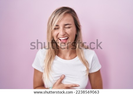 Young blonde woman standing over pink background smiling and laughing hard out loud because funny crazy joke with hands on body. 