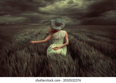 Young blonde woman standing in a wheat field, wearing a yellow summer dress, on a windy and stormy day