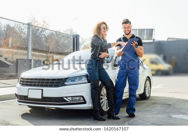 Young blonde woman\
stands with a man who washes cars at the self-service, he holding\
blue folder and smile.