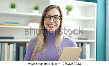 Young blonde woman student smiling confident holding clipboard at library university