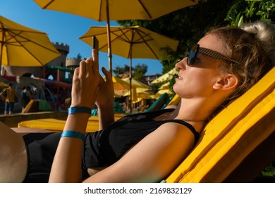 Young Blonde Woman In Sunglasses In A Black Swimsuit Is Lying On A Yellow Sun Lounger With An Umbrella By The Pool With A Mobile Phone In Her Hands. Side View, In The Sunset Light