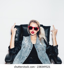 Young blonde woman in sunglasses sit on black office chair and showing middle finger . Fashion hipster girl.  White background, not isolated