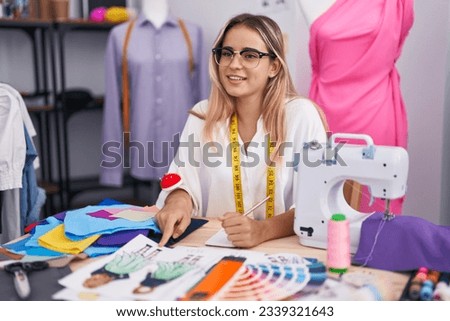 Young blonde woman tailor pointing to clothing design speaking at clothing shop