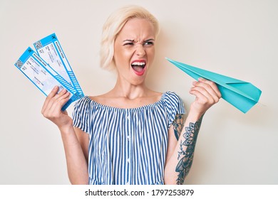 Young Blonde Woman With Tattoo Holding Paper Airplane And Boarding Pass Angry And Mad Screaming Frustrated And Furious, Shouting With Anger. Rage And Aggressive Concept. 