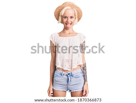 Young blonde woman with tattoo wearing summer hat sticking tongue out happy with funny expression. emotion concept. 